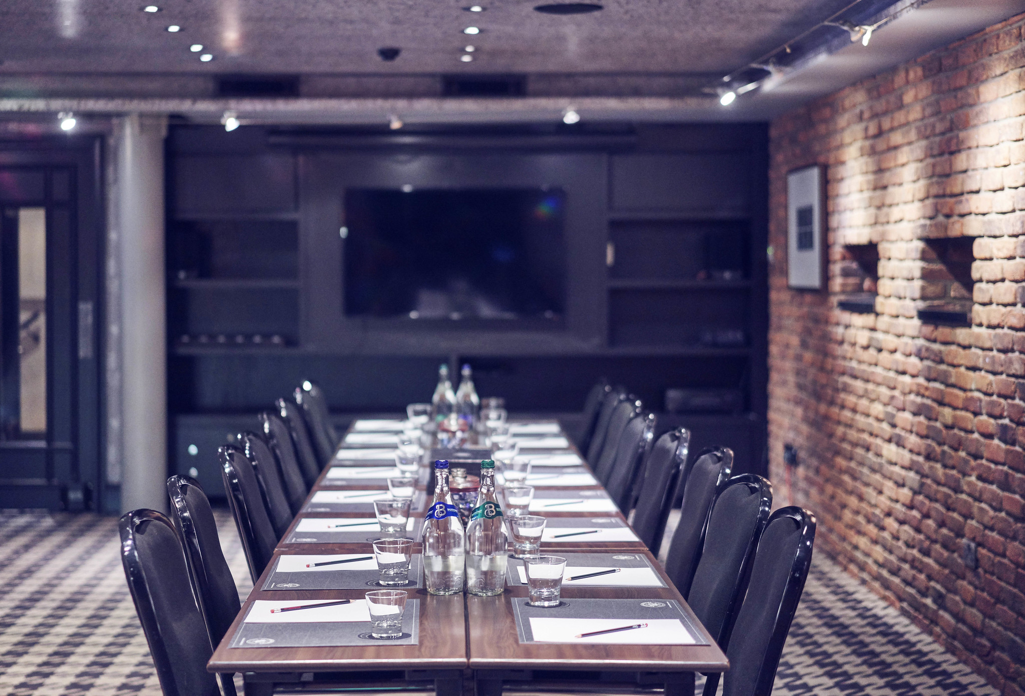 View of a meeting table with 16 black chairs on a black and white carpet, an exposed brick wall with artwork is to the right of the table, and a flat screen TV can be seen in the background.